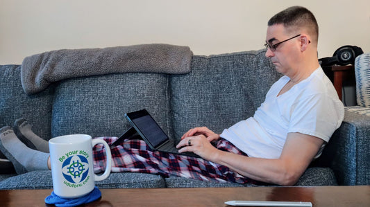 The author, wearing pajamas, is reclining on a couch and typing on a small laptop.