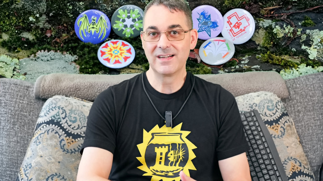 Martin is sitting on his couch wearing a black T-shirt featuring Skeltofish, a skeletal fish in a skeletal-form fishbowl with a gothic tower tchotchke. The wall behind him is a picture of a mossy rock with buttons featuring more of his designs.