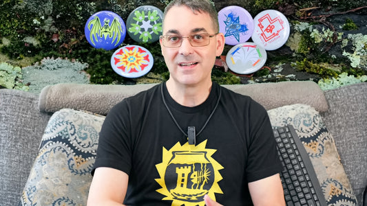 Martin is sitting on his couch wearing a black T-shirt featuring Skeltofish, a skeletal fish in a skeletal-form fishbowl with a gothic tower tchotchke. The wall behind him is a picture of a mossy rock with buttons featuring more of his designs.